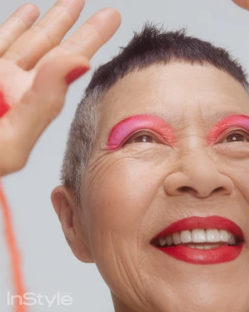 Close-up portrait of an elderly woman with bright pink eye makeup and red lipstick smiling with hand raised, by Nicole Pinky Thompson